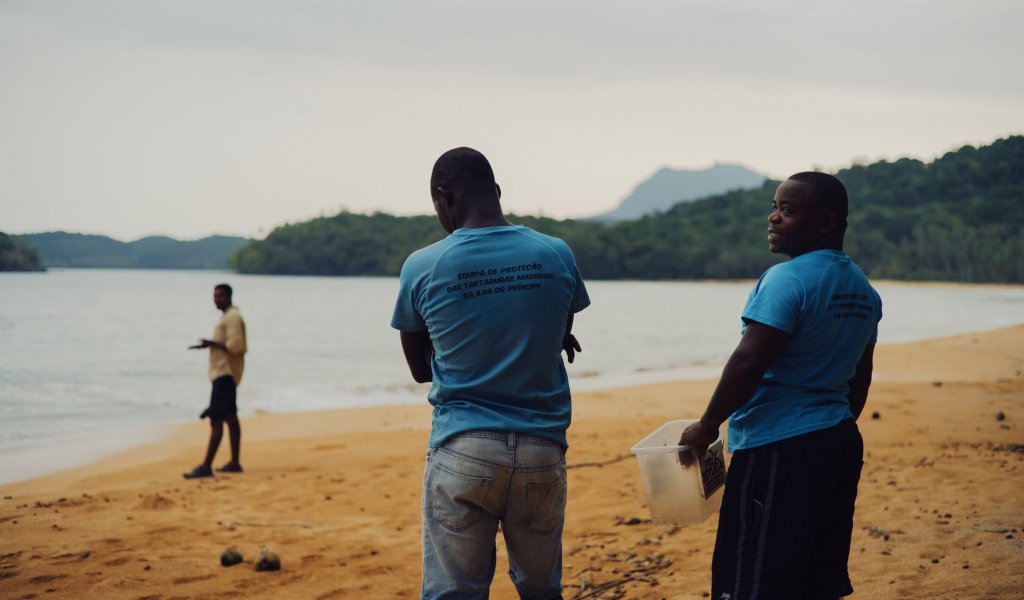 Three locals on Principe Island beach, one holding a plastic container.