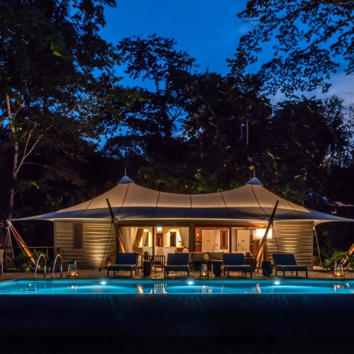 Vista exterior da tenda-vila no Sundy Praia à noite, com o quarto iluminado ao fundo e a piscina em primeiro plano.