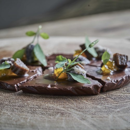 Close-up de uma elegante sobremesa de chocolate decorada com ervas e frutas, servida num prato de madeira no restaurante OKA Sundy