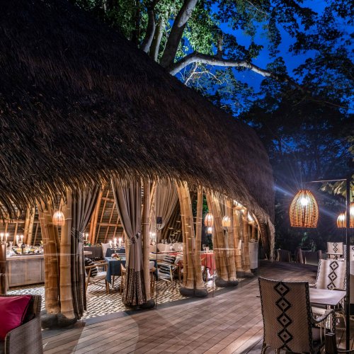 Área de estar ao ar livre e estrutura de bambu do restaurante OKA Sundy rodeada por vegetação densa