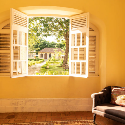 Janela iluminada no Belo Monte com vista para os jardins do pátio e edifícios amarelos, poltrona acolhedora no interior.