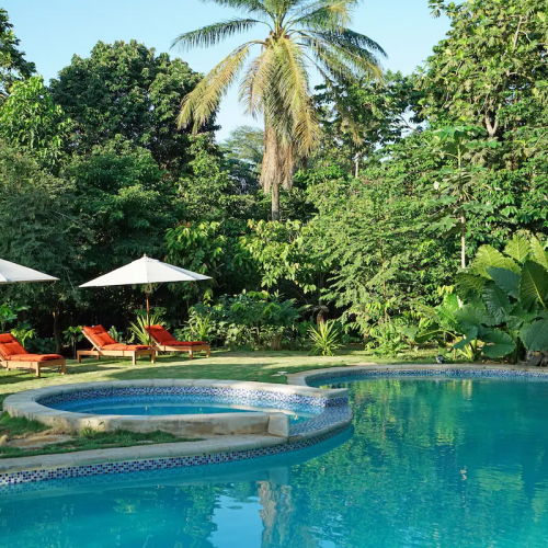 Curved swimming pool at Belo Monte, red loungers and white umbrellas in a lush garden.