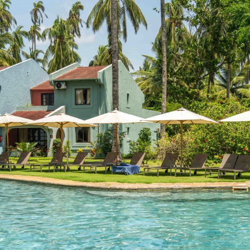 Espreguiçadeiras e chapéus de sol junto à piscina do Omali, com edifícios em tons pastel e palmeiras ao fundo, Ilha de São Tomé