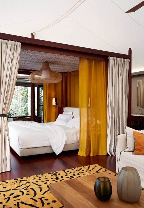 View of the elegant double bed in a tent villa at Sundy Praia, with a wooden table, vases, and a yellow-black rug in the foreground.