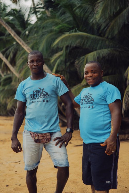 Dois locais com camisetas Pro Tetuga azul claro e calções sorriem para nós na praia da Ilha do Príncipe.
