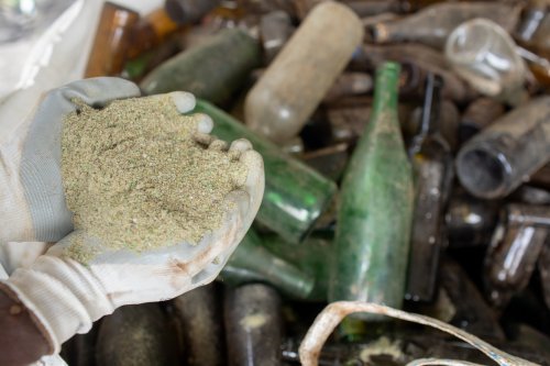 Worker's hand holding glass sand, with many empty glass bottles in the background.