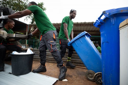 Três trabalhadores com camisetas verdes a reciclar com contentores azuis na Ilha do Príncipe.