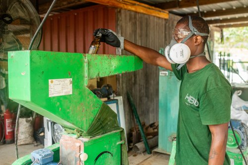 Trabalhador com T-shirt verde escuro HBD, máscara e óculos de proteção a reciclar vidro com uma máquina na Ilha do Príncipe.