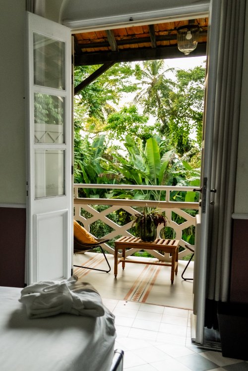 View from a bedroom at Roça Sundy showing a balcony with a chair, table, and lush greenery