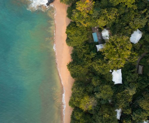 Vista aérea das tenda-vilas no Sundy Praia, quase escondidas pela selva, com a praia e a água cristalina à frente.