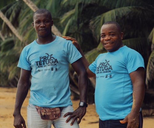 Two locals in light blue Pro Tetuga T-shirts and shorts smile at us on Príncipe Island beach.