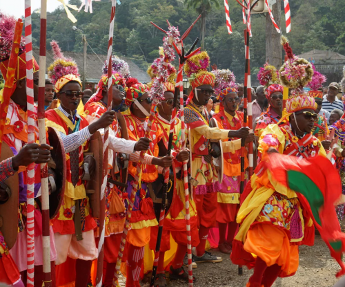 Desfile carnavalesco no Príncipe, figurantes em trajes tradicionais coloridos com bandeiras, varas e tambores.