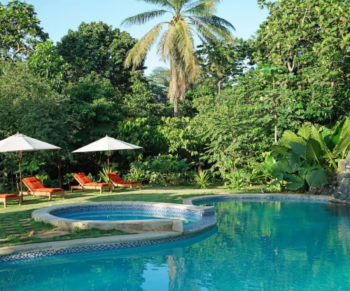 Curved swimming pool at Belo Monte, red loungers and white umbrellas in a lush garden.