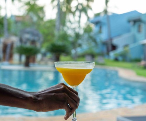 Mão a segurar um cocktail tropical junto à piscina do Omali, com palmeiras e edifícios ao fundo, Ilha de São Tomé