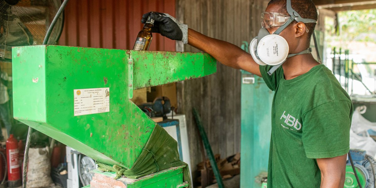Trabalhador com T-shirt verde escuro HBD, máscara e óculos de proteção a reciclar vidro com uma máquina na Ilha do Príncipe.