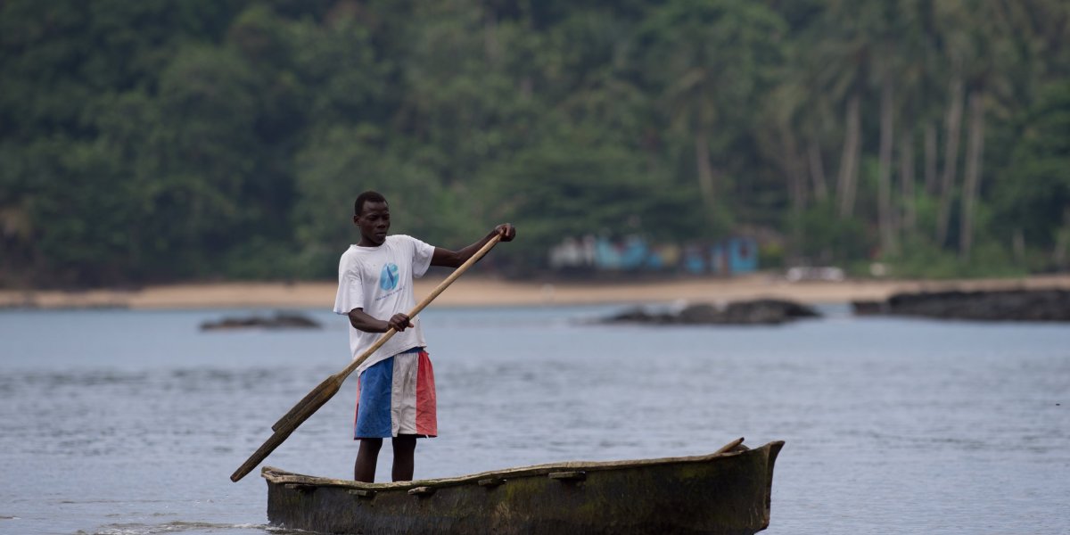 A local fisherman in colourful trousers rowing a small wooden boat, with the beach and rainforest in the background.