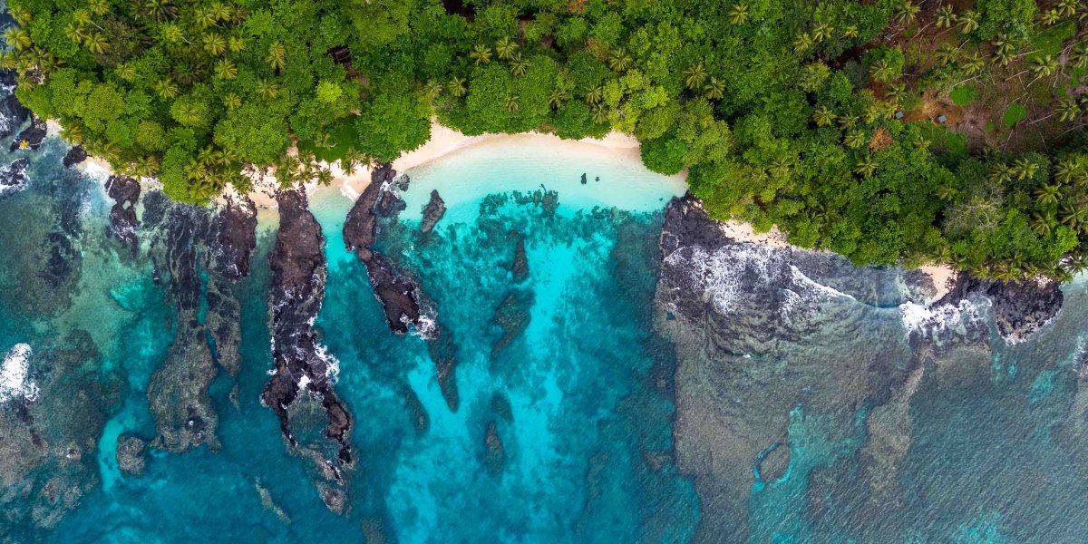 Vista aérea de uma praia na ilha do Príncipe, com floresta tropical, areia branca e águas cristalinas sobre formações vulcânicas