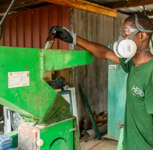 Trabalhador com T-shirt verde escuro HBD, máscara e óculos de proteção a reciclar vidro com uma máquina na Ilha do Príncipe.