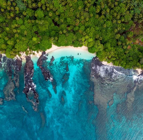 Vista aérea de uma praia na ilha do Príncipe, com floresta tropical, areia branca e águas cristalinas sobre formações vulcânicas
