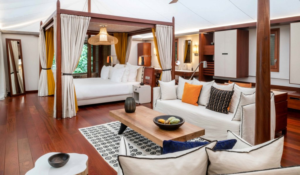 Quarto com cama de dossel elegante, tapete artístico, área de estar e mesa de madeira em primeiro plano na tenda-vila Sundy Praia.
