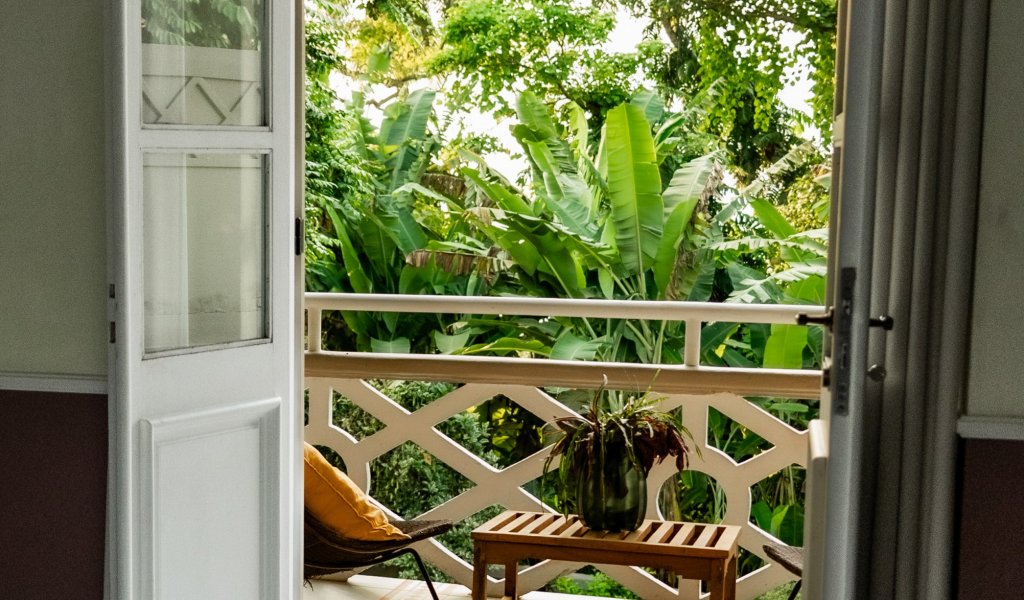 View from a bedroom at Roça Sundy showing a balcony with a chair, table, and lush greenery