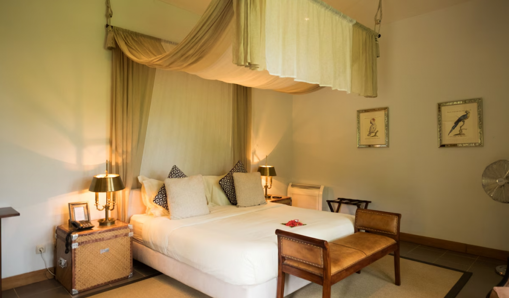 Panoramic Suite bedroom with canopy bed, warm lamps and leather bench, framed bird prints on the wall.