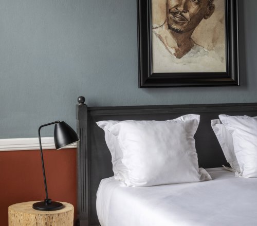 Bedroom detail at Roça Sundy's Eclipse Yard Room, with a black bed frame, white linen, a wooden stump bedside table and a framed watercolour portrait on a blue-grey wall, São Tomé and Príncipe