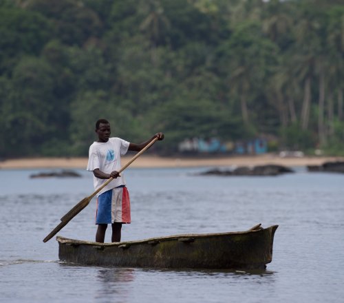 A local fisherman in colourful trousers rowing a small wooden boat, with the beach and rainforest in the background.