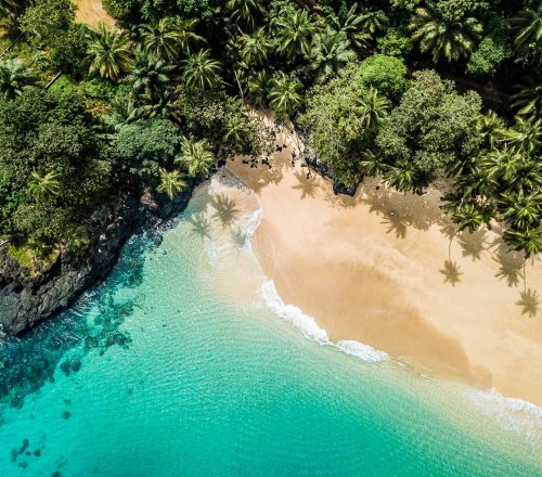 Vista aérea de uma praia isolada na Ilha do Príncipe, com areia dourada, águas turquesa e rochas vulcânicas emolduradas por coqueiros